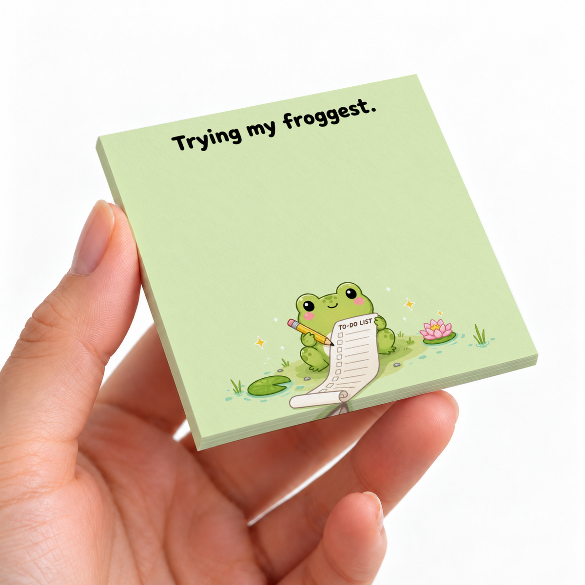 Hand holding a green frog sticky note pad with “Trying my froggest” text and a frog writing a to-do list.