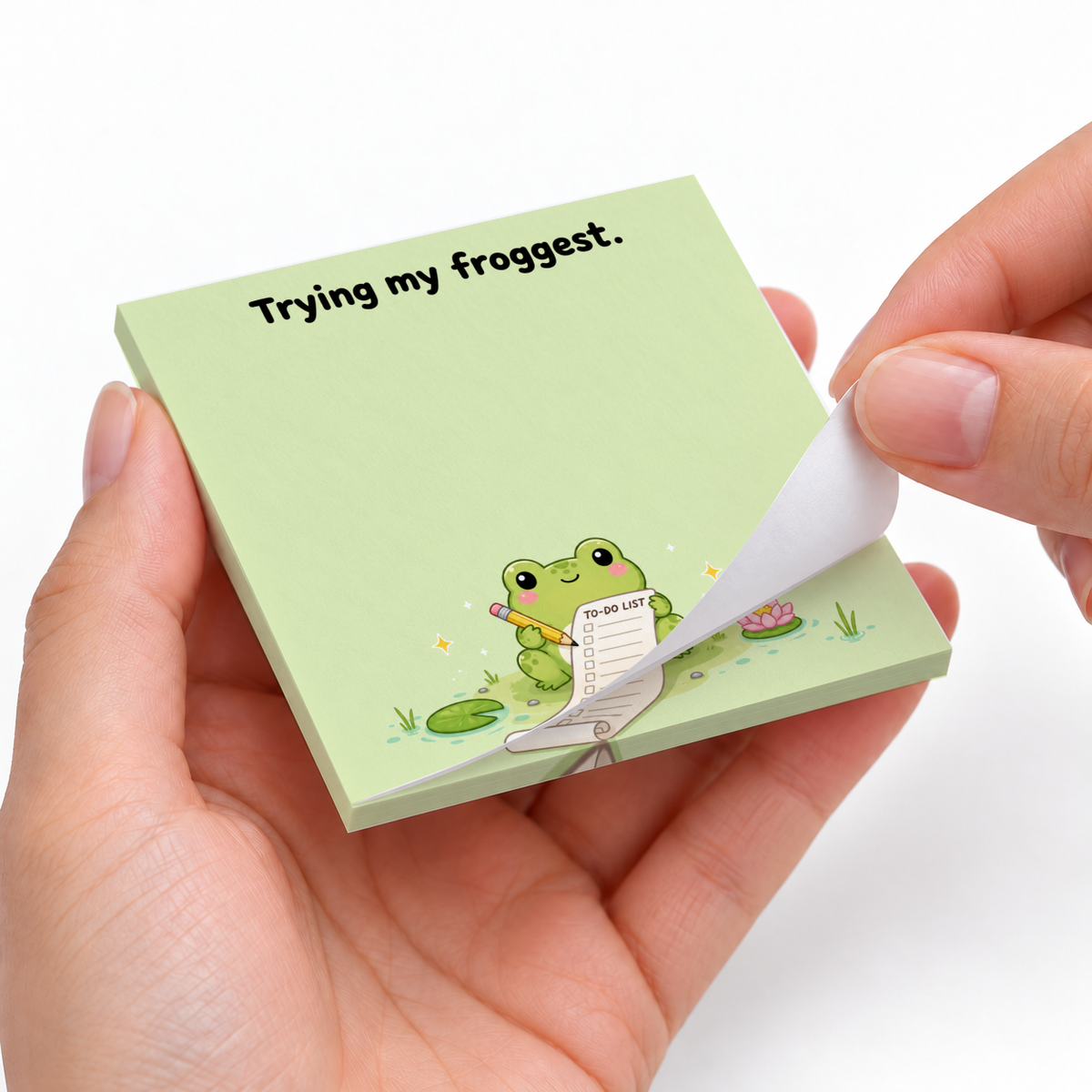Hands peel a sheet from the green “Trying my froggest” frog sticky note pad with a frog holding a to-do list.