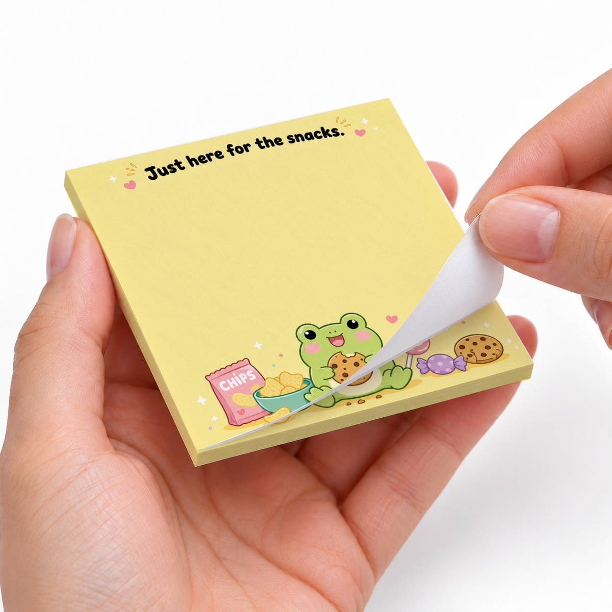 Hands peel a sheet from a yellow frog and snacks sticky note pad reading “Just here for the snacks.”