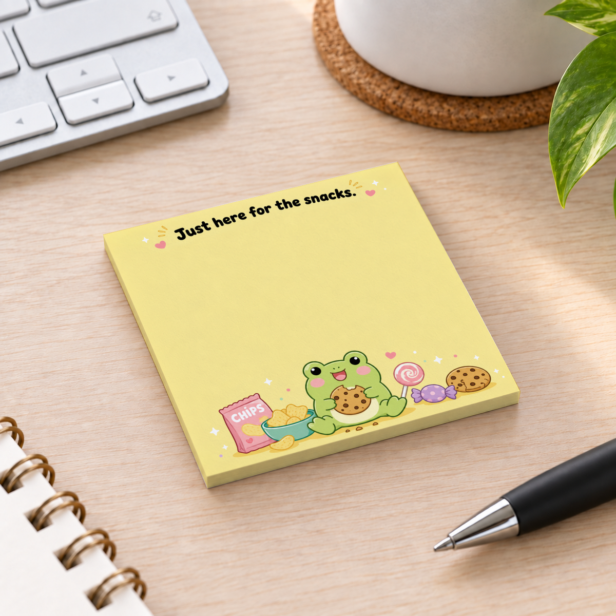 Yellow frog and snacks sticky note pad on a wooden desk beside a keyboard, pen, notebook, and potted plant.