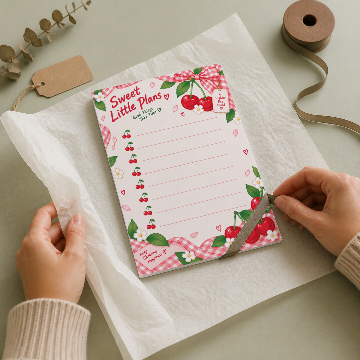 Hands wrapping a cherry tear-off memo pad in white tissue paper on a light green desk with ribbon and a blank tag.