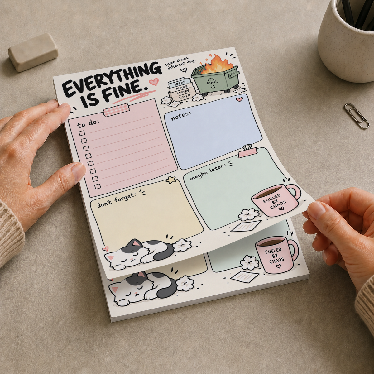 Hands tearing a sheet off the Everything Is Fine Dumpster Fire Memo Pad on a desk with office supplies.
