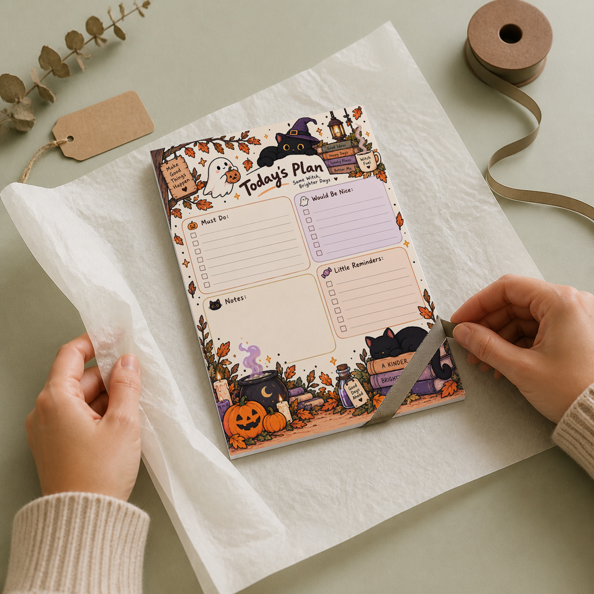 Hands unwrapping a Spooky Witch Cat Today's Plan Notepad from tissue paper on a desk with a ribbon spool and gift tag.