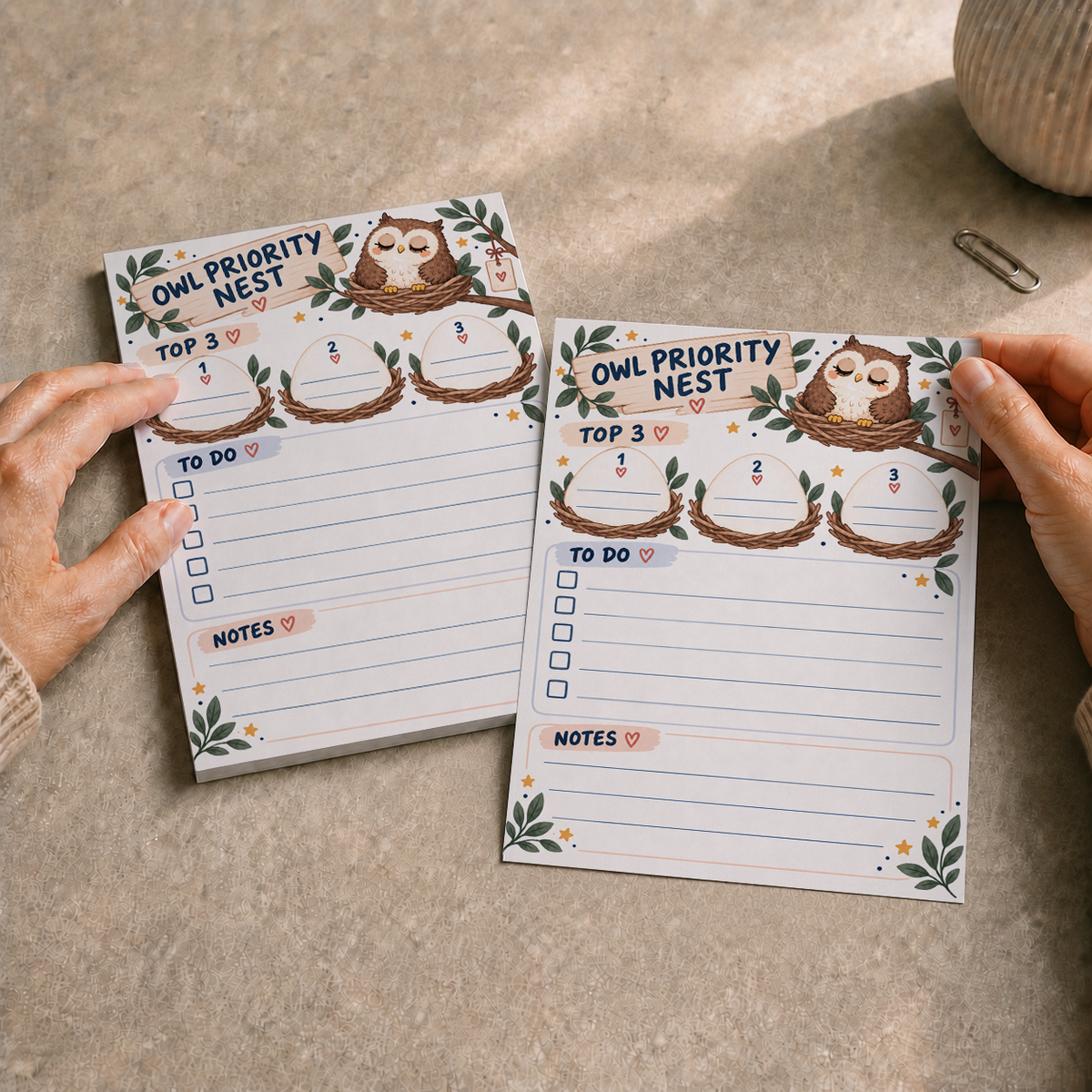 Hands holding a sheet from the Owl Priority Nest Notepad next to the full tear-off memo pad on a textured desk.