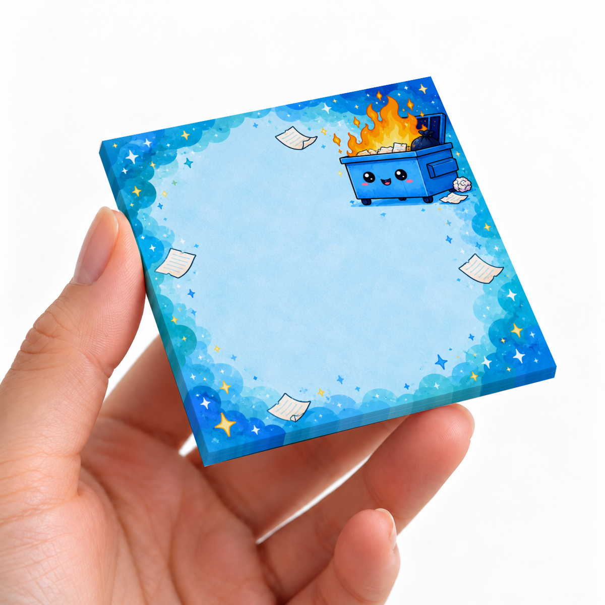 A hand holds a blue sticky note pad featuring a smiling dumpster fire, scattered papers, and a starry border.