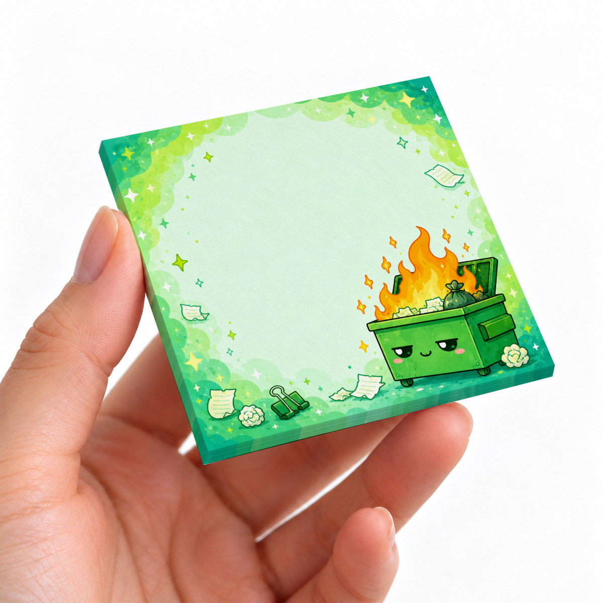 Hand holding a kawaii green dumpster fire sticky note pad with a blank center against a white background.