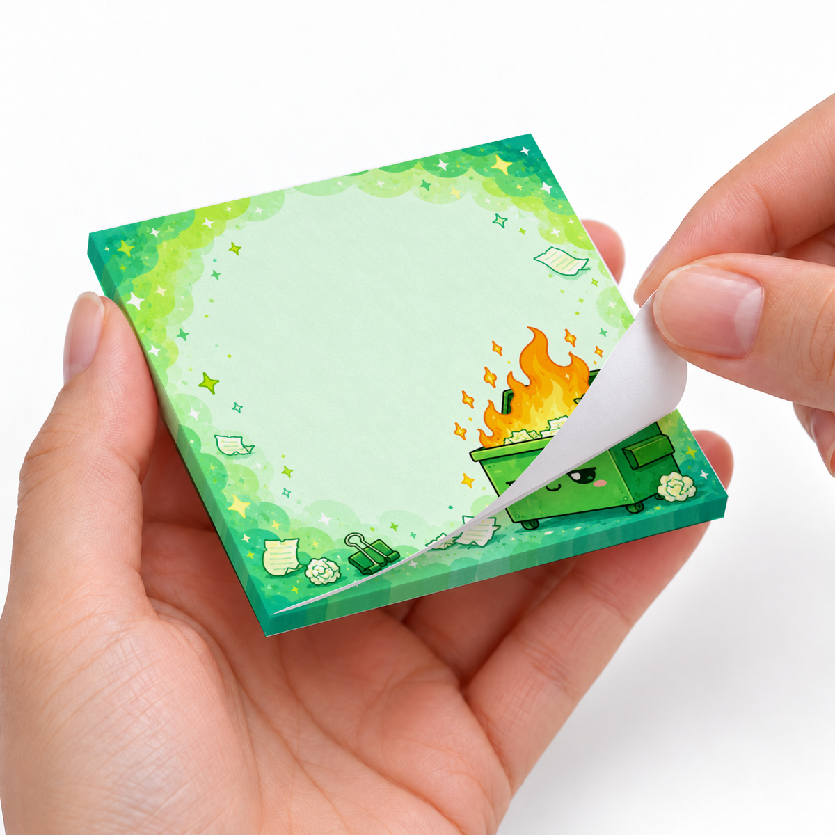 Hands peel a sheet from the kawaii green dumpster fire sticky note pad against a white background.