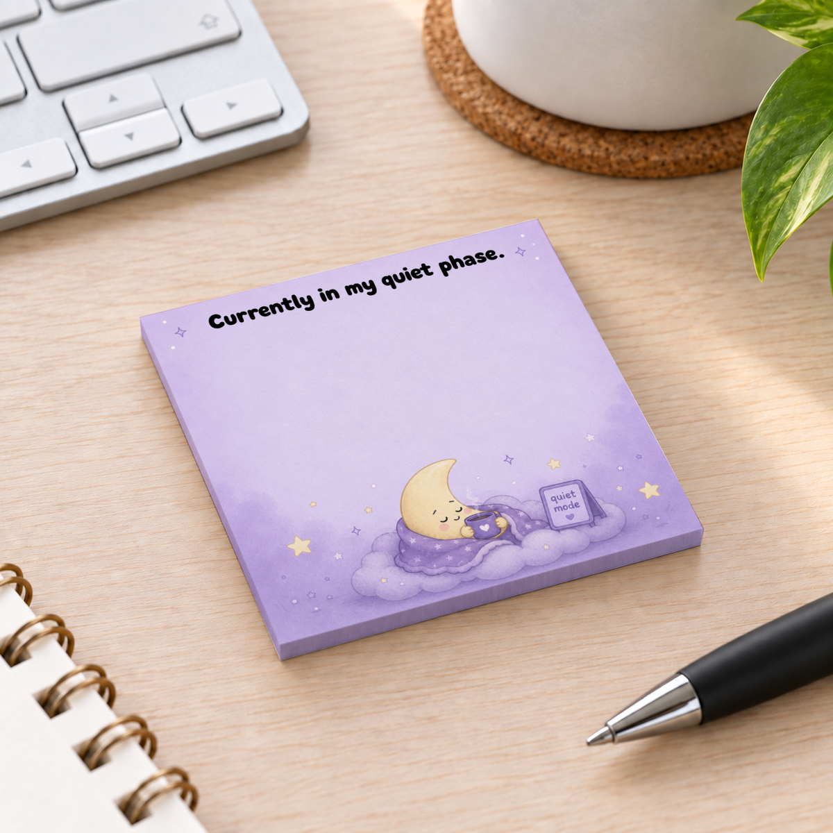 Purple “Currently in my quiet phase” moon sticky note pad on a desk beside a keyboard, notebook, pen, and plant.