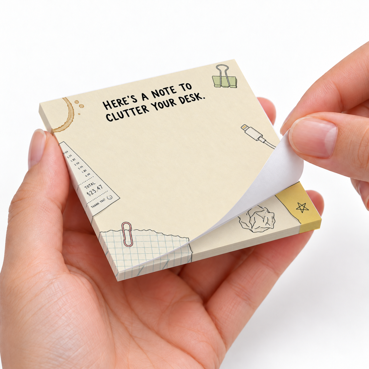 Hands peel a sheet from the Desk Clutter Doodles sticky note pad with office-themed illustrations.
